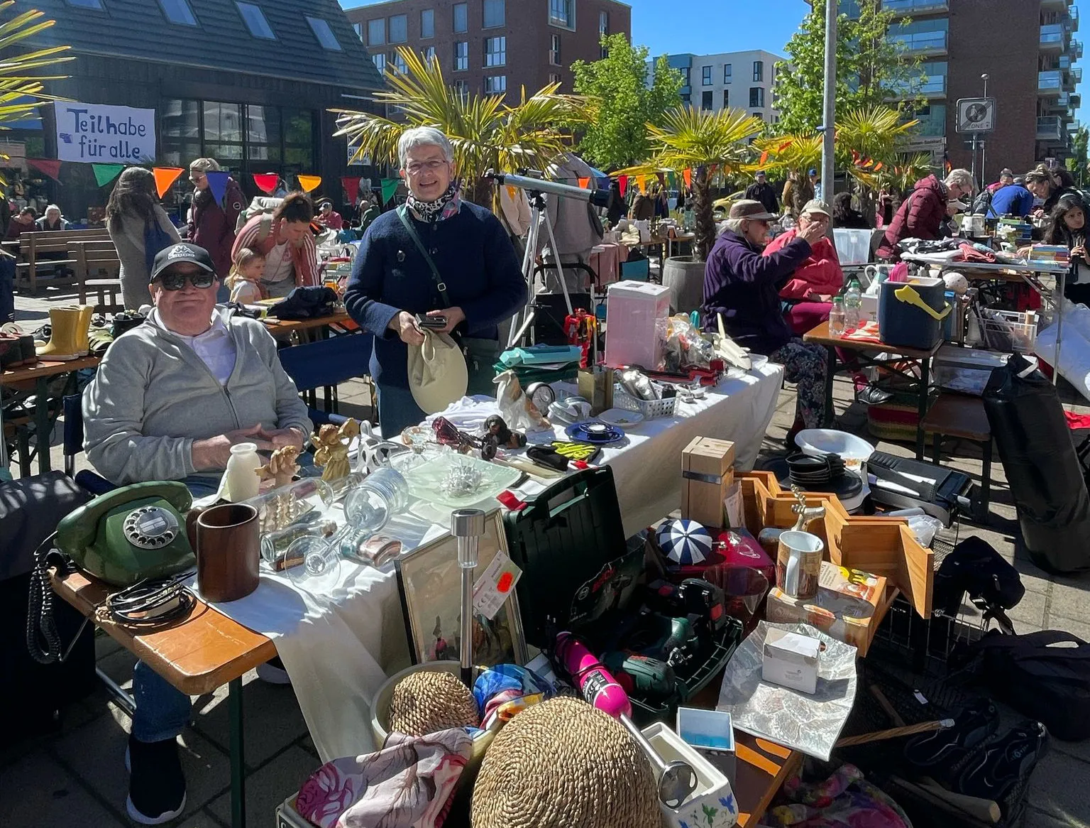 Zwei Personen an einem Flohmarktstand schauen lächelnd in die Kamera. Der Flohmarktstand ist voll mit unterschiedlichsten Sachen, darunter ein Telefon mit Drehscheibe. Rechts davon befinden sich weitere Flomarktstände und Personen. Dahinter sind Bäume, Wimpelketten und Gebäude zu sehen.