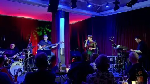 Eine Sängerin hält ein Mikrofon und singt, links von ihr spielt ein Bassist. Publikum schaut der Band zu.