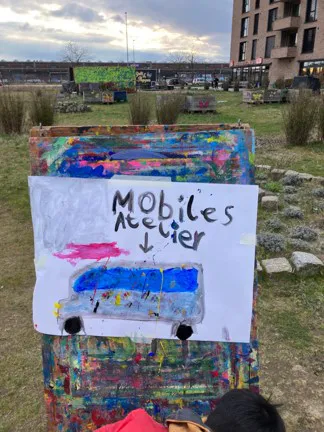 Auf einer Wiese steht eine Staffelei. Darauf ist ein großes Blatt Papier im Querformat. Auf dem Blatt ist ein Bulli gemalt, darüber steht Mobiles Atelier. Ein Pfeil zeigt von der Schrift auf den Bulli.