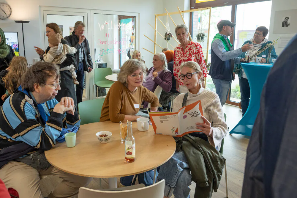 An einem runden tische sitzen drei Personen. Eine blättert in einer Broschüre, die zweite schaut mit ins Heft. Im Hintergrund sind weitere Personen am stehen und im Gespräch.