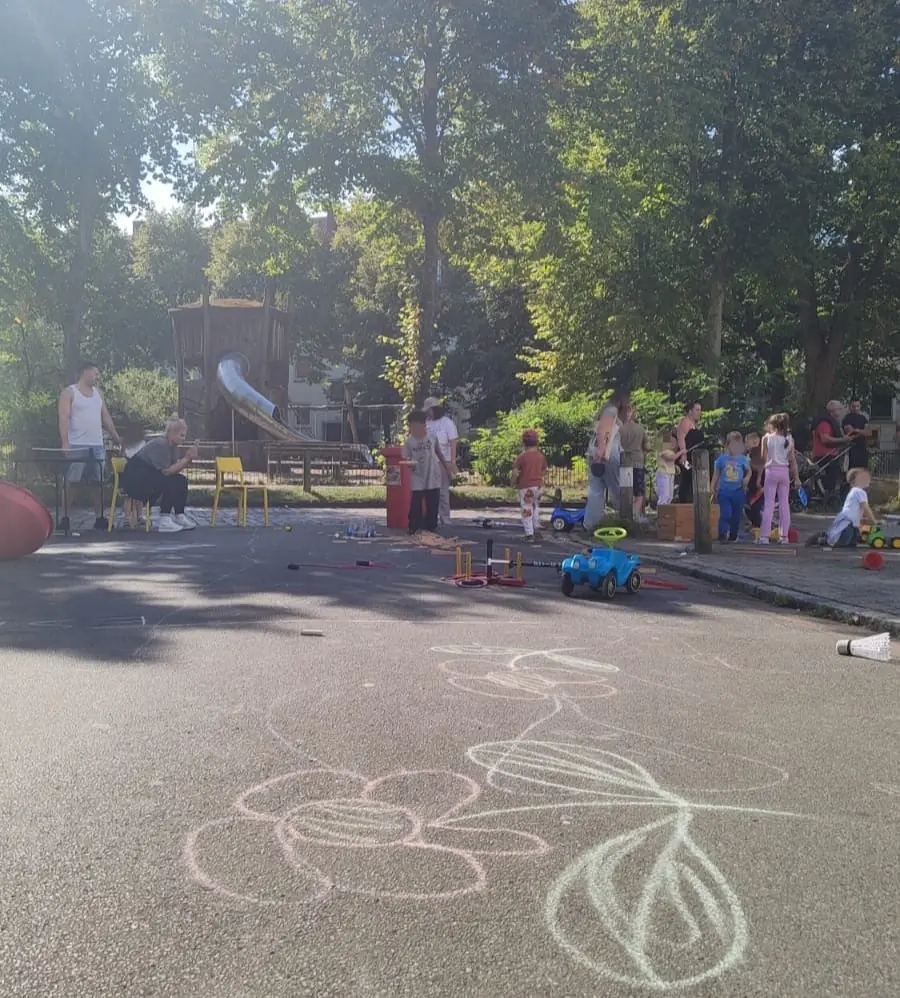 Kinder und Erwachsene spilen bei einem Straßenfest auf der Straße. Auf den Asphlt wurde mit Kreide eine Blume gemalt. Im Hinterrund sind Bäume und ein Spielplatz zu sehen.
