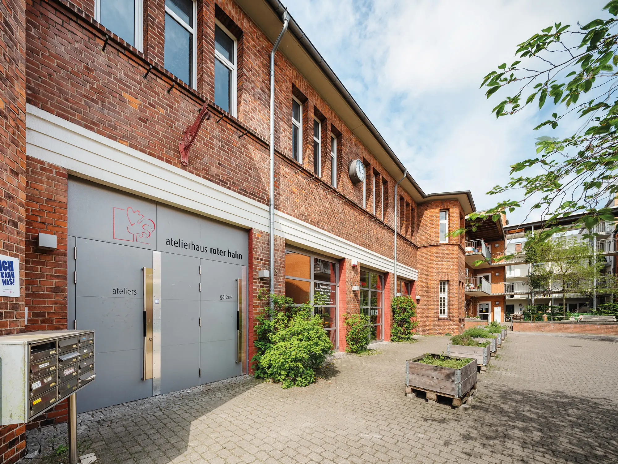 Ein Backsteingebäude mit mehreren Fenstern und grauen Türen. Auf den Türen steht „atelierhaus roter hahn“. Davor stehen Pflanzkübel, Briefkästen und einige Büsche.