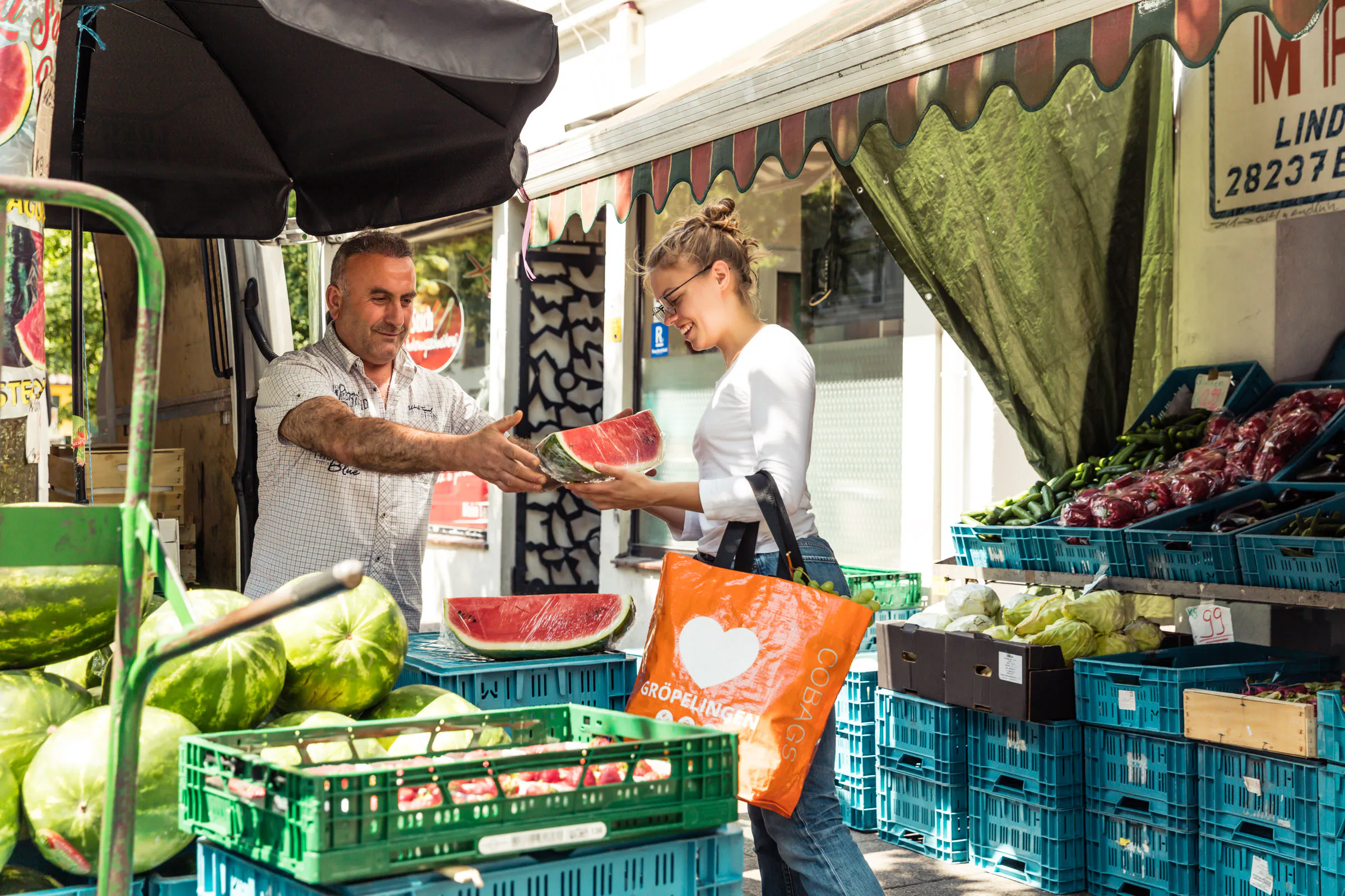 Ein Gemüsehändler reicht der Kundin ein Stück Wassermelone. sie hat einen Fahrradtasche überm Arm hängen auf der ein Herz zu sehen ist.