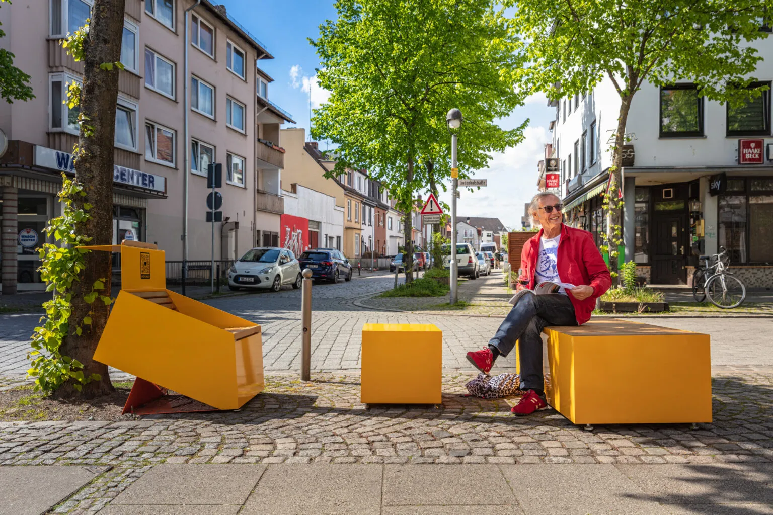 Auf dem Bild sitzt ein Mann auf einer knallgelben Sitzbank mitten auf einem kleinen Platz in der Stadt. Um ihn herum stehen weitere gelbe Möbel – ein Hocker und eine schräge Liegefläche. Es ist sonnig, die Bäume spenden etwas Schatten, und der Mann trägt ein auffälliges rotes Outfit. Die Szene wirkt entspannt und offen – als würde dieser Ort zum Verweilen und Plaudern einladen.