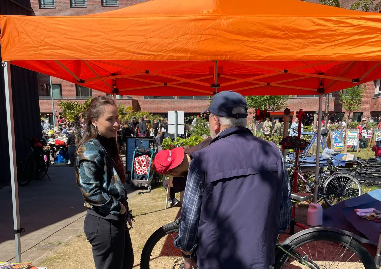 Zwei Personen befinden sich im Gespräch. Eine weitere Person reparier sein Fahrrad. Dahinter steht ein Pavillon.