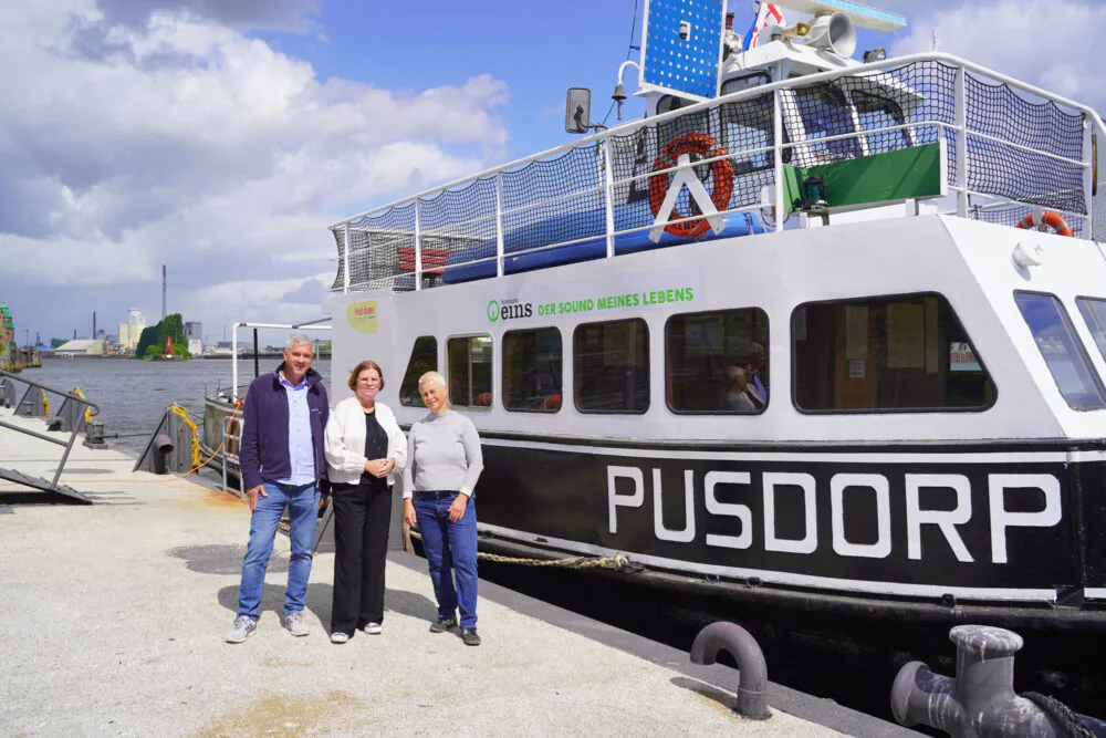 Auf einem Schwimmanleger stehen drei Personen und schauen in die Kamera. Am Anleger liegt ein Fahrgastschiff mit dem Namen "Pusdorf" der Name ist zu sehen. Im Hintergrund befindet sich eine Rampe mit Geländer, Wasser und ein bewölkter Himmel.