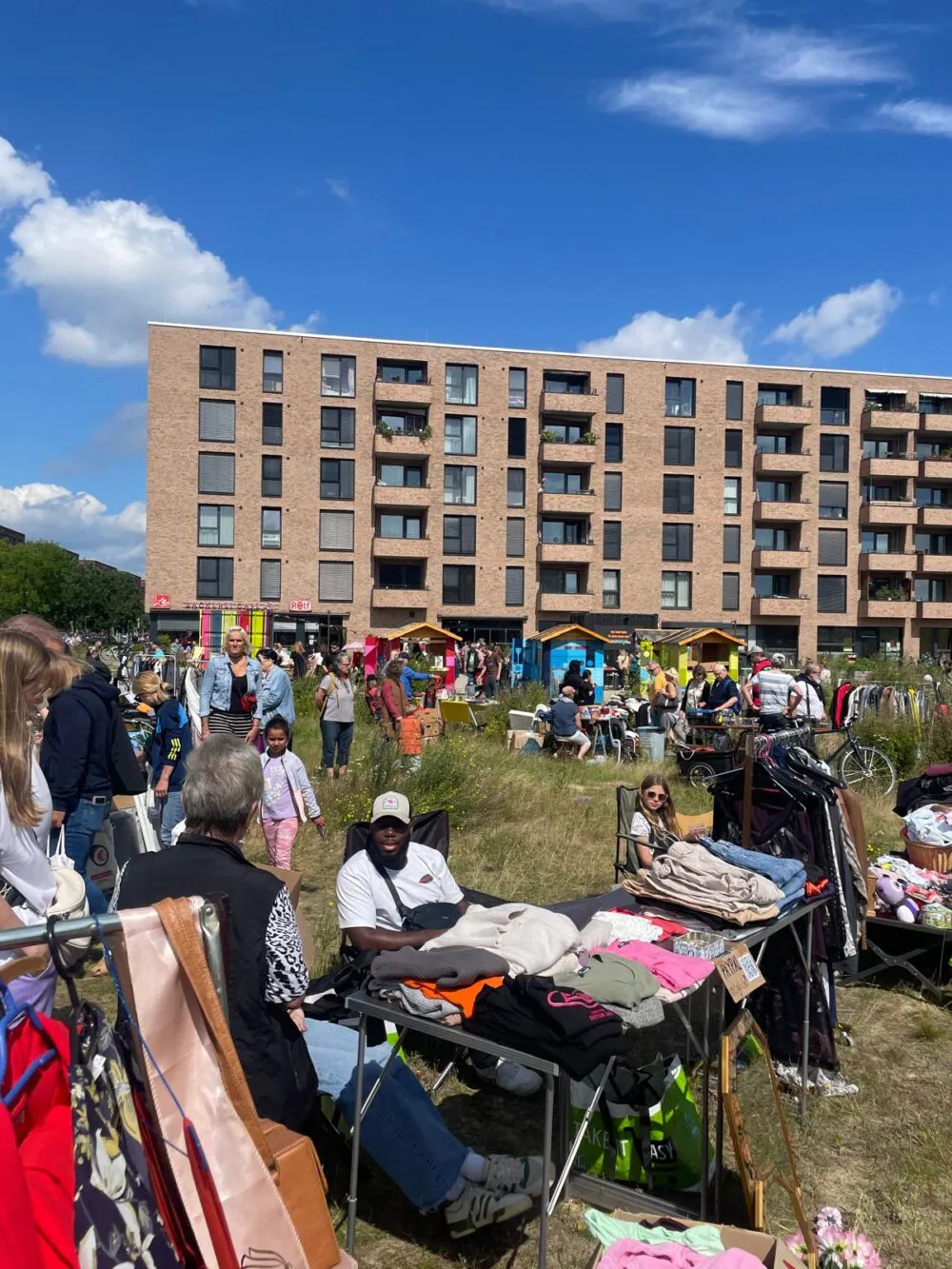 Auf einer Wiese vor einem großen Gebäude sitzen und stehen viele Personen. Ein Mensch ist im Vordergrund zu sehen. Sie sitzt hinter einem Tisch mit auf dem viele Klamotten liegen - auf der Wiese findet ein Flohmarkt statt.