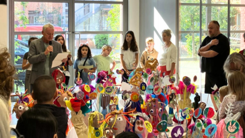 In der Galerie im Atelierhaus Roter Hahn wird eine Ausstellung eröffnet. Kinder und Erwachsene hören einem Redner zu. In der Mitte zwischen den Personen sind Skulpturen und Plastiken zu sehen.