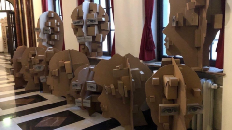 Große skulpturale Köpfe aus Pappe stehen nebeneinander im Flur eines Gebäudes mit Marmorboden und hohen Fenstern. Die geometrisch aufgebauten Gesichter sind mit aufgeklebten Augen versehen.