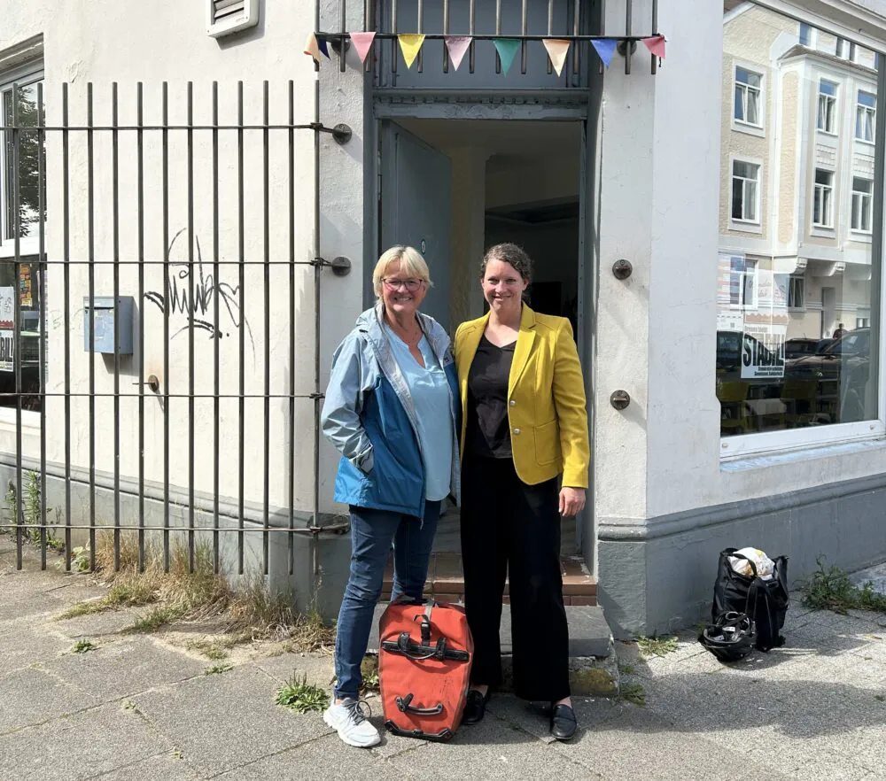 Vor einem geöffneten Ladenlokal stehen zwei Personen dicht beieinander und lächeln in die Kamera.